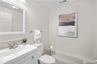 Remodeled in 2026, the lower level bath reflects the same fresh, cohesive tone found throughout the home. Soft neutral paint complements the new flooring, creating a clean and welcoming backdrop.    The new vanity is topped with a smooth white cultured marble counter that flows gently into the sink, giving the space a seamless, polished look. A band of small mosaic tiles along the backsplash adds quiet texture behind the faucet. The new toilet and brushed nickel finishes coordinate beautifully, echoed in the vanity light above where frosted shades cast a bright, uplifting glow.