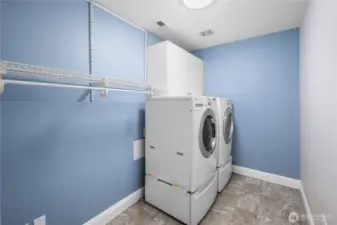 The lower level laundry room is generously sized, giving you plenty of room to move around and keep things organized. A full front-load washer and matching dryer sit side by side, ready for everyday loads or larger laundry days.  Open shelving and hanging space stretch along the wall, offering room for baskets, linens, and sorting stations, while still leaving floor space for a folding table or additional storage if you choose. Bright paint tones keep the space feeling cheerful and clean, turning a practical task into a pleasant part of the day.