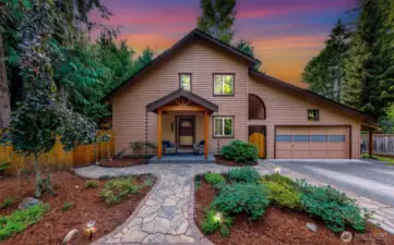 At twilight, the front of the home glows against a vibrant sky, with uplighting illuminating the landscaped entry and charming covered porch. A stunning introduction to this private Geneva retreat.