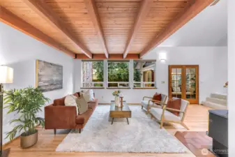 This view of the living area highlights the flow of natural light and the home’s connection to nature through its picture windows and forested backdrop. The French doors invite movement between living spaces and the sunroom beyond.