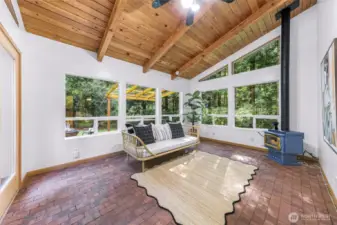 The sunroom is a bright, tranquil haven, featuring vaulted ceilings, exposed beams, and a freestanding wood stove. Oversized windows capture peaceful views of the wooded backyard, blurring the line between indoor and outdoor living.