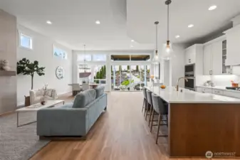 Straight-on view of the open-concept living space with the living room to the left, designer kitchen to the right, and a dramatic wall of windows framing the landscaped backyard — showcasing light-filled newer construction living in McCormick Trails, Port Orchard.
