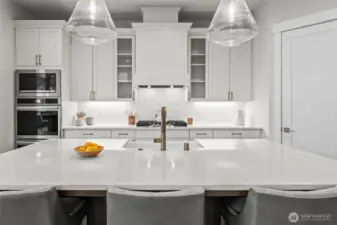Straight-on view from the kitchen island highlighting custom cabinetry, quartz countertops, specialty lighting, and stainless steel appliances, designed for gathering and everyday living within the open-concept layout of this upgraded newer construction home in McCormick Trails, Port Orchard.