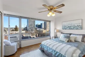 The bedroom keeps the same bright, open feel with expansive windows, a generous mirrored closet that amplifies the space, and a window A/C unit for warm days.