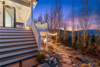 Twilight view of the home's outdoor entertaining area w/ stairs to the main level view deck & pavers to the private lower patio w/ firepit & hot tub w/ west-facing uplake views of Lake Chelan.