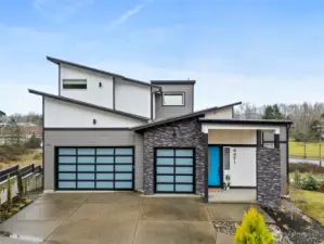 Modern exterior of a contemporary home for sale in the River Rock subdivision, featuring clean architectural lines, stone accents, a spacious driveway, and attached garage. Aerial and front views highlight its setting within the scenic River Rock community surrounded by natural greenery.