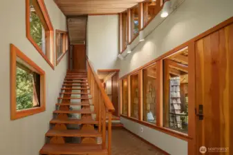 Open wood stairs rise through a dramatic two-story hall, guiding you to the primary suite and a hallway with two additional bedrooms. A door off the hall opens to a private deck—perfect for enjoying the surrounding nature—while windows on both sides fill the space with light.