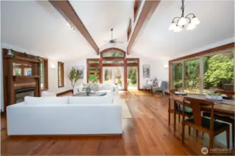 Stunning great room with soaring vaulted ceilings, exposed wood beams, and expansive windows that bring in natural light and serene outdoor views. The open-concept layout seamlessly connects the living and dining areas, centered around a cozy fireplace and framed by rich wood finishes throughout. Large sliding doors provide effortless indoor-outdoor living, creating an inviting space perfect for relaxing or entertaining.