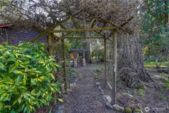 Charming garden pathway framed by a rustic wood arbor, creating a peaceful, storybook entrance through the lush landscape. Mature trees, natural stone accents, and layered greenery offer a tranquil setting that feels private and inviting.