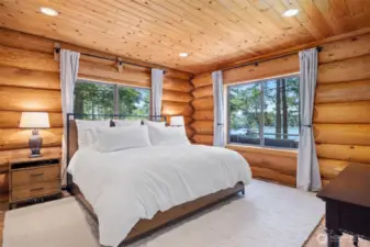 Main floor primary bedroom offers a tranquil retreat with dual picture windows capturing water views, log walls, and natural textures that blend luxury with comfort.