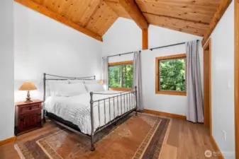 Vaulted cedar ceilings and large windows create a bright yet cozy retreat in the upper level guest room. The peaceful forest view, hardwood floors, and clean neutral palette offer a relaxing space to unwind.