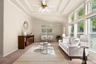 The living room feels bright, open, and inviting. Large picture windows line one wall, filling the space with natural light and offering views of the greenery outside. A tray ceiling with crown accents and a ceiling fan adds an elegant touch and a sense of height.