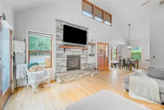 Open-concept living and dining space featuring vaulted ceilings, a floor-to-ceiling stone fireplace with mounted TV, and abundant natural light—perfect for modern, comfortable living.