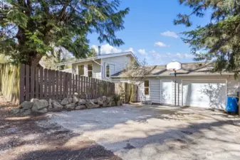 The driveway side: rock retaining wall, attached garage, and a basketball hoop that conveys. Mature trees on a fully fenced corner lot.