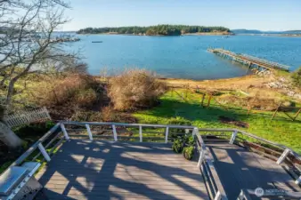 View across Fisherman Bay from main house, upper level deck (observation deck), looking down on main level deck, fenced garden, and then fire pit and picnic area on the beach. The peninsula and Spit Preserve (Lopez Island) seen across the water, with the entrance to Fisherman Bay off to the right