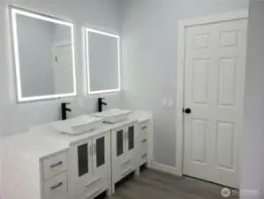 A Full View of the Primary Master Bath Room Showing New Double Sink Vanity, Sinks and Faucet, Two Lighted Mirrors, New Toilet, Paint, Trim and Vinyl Plank Flooring.