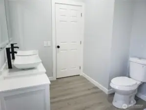 A View of the Primary Master Bath Room Showing New Double Sink Vanity, Sinks and Faucet, Two Lighted Mirrors, New Toilet, Paint, Trim and Vinyl Plank Flooring.