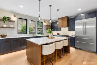 The central island anchors the kitchen with its sleek white countertop and warm wooden base, offering both a stylish prep space and a casual spot for gathering. Nearby, the stainless steel refrigerator adds a bold, modern touch with its clean lines and spacious double-door design, perfectly complementing the kitchen’s upscale aesthetic.