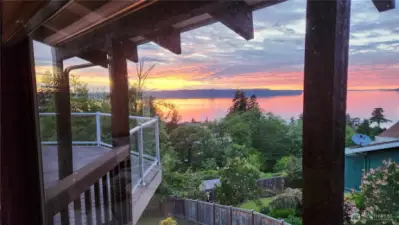 Breathtaking sunset views from the comfort of your own home. This stunning vantage point captures the vibrant colors over Puget Sound, framed by the deck’s elegant glass railing and natural surroundings.