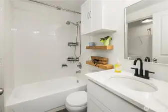 Bathroom with quartz countertops and black durable matte finish faucets. Plenty of storage space in cabinets and wooden, thick shelves. Bath tub with shower products holder in it. Bathroom connected with laundry room.