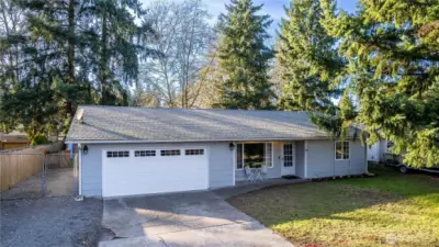 This front exterior view highlights the home’s welcoming curb appeal and well-maintained presence from the street. The wide driveway offers ample parking and leads to a spacious two-car garage, providing both convenience and additional storage options. The home’s low-profile roofline and classic rambler design create an approachable, easy-living aesthetic, while the siding appears clean and cared for. Mature trees frame the property, adding privacy, shade and a natural Pacific Northwest backdrop that enhances the overall setting. The front lawn provides usable green space without high maintenance demands, and the covered entry offers protection from the elements while creating a defined arrival point. Altogether, the exterior presents a move-in ready first impression that feels comfortable, established and inviting the moment you pull up.