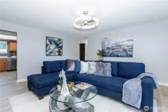 The main living area opens up into a spacious, comfortable setting centered around a classic fireplace that acts as the room’s focal point. The white brick surround adds texture and character without feeling dated, blending nicely with the updated flooring and fresh paint tones. Large windows bring in soft natural light, helping the room feel bright without being harsh. The layout easily accommodates a full sectional while still leaving breathing room, making it ideal for both entertaining and everyday lounging. It’s a practical, livable space that balances warmth and openness.