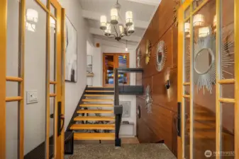 View of the foyer with its double glass doors, striking wood paneled wall and architectural staircase.