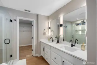 Primary bathroom featuring a wide dual vanity with large mirrors and modern wall lighting that brightens the entire space. The layout flows toward a spacious walk-in shower and adjacent walk-in closet, creating a comfortable and practical daily routine space.
