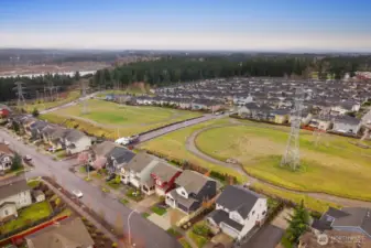 Expanded aerial view capturing the surrounding homes, open fields, and nearby forested landscape. The elevated perspective showcases the balance of neighborhood living and natural surroundings.