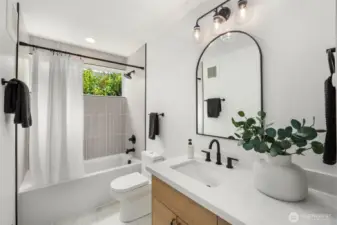 Fresh, modern style in the main floor bathroom with a frameless arched mirror, stylish vanity lights, vertical tile surround, and a deep soaking tub paired with a shower, set beneath a bright window.