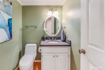 The main-level powder room features a modern vanity with a dark stone countertop and a fresh, stylish color scheme. Thoughtful lighting and a round mirror enhance the space, making it both functional and aesthetically pleasing.