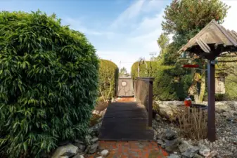 A small wooden bridge leads through the garden to an artistic gate, creating a welcoming approach that feels both whimsical and serene.