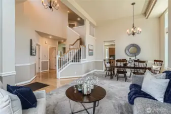 This stunning interior showcases a spacious open-concept layout with soaring ceilings and elegant architectural details. A grand staircase with white spindles and wooden handrails serves as a striking focal point, leading to the upper level. Warm hardwood flooring flows through the entryway, complementing the neutral color palette and crisp white trim. The inviting living and dining areas are carpeted for comfort, with large windows flooding the space with natural light. A stylish chandelier adds charm above the formal dining area, while a cozy sitting area with plush seating and a sleek coffee table completes this beautifully designed, welcoming space.