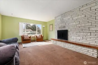 The first of two large living spaces, the living room features a brick fireplace and hearth, and bright sunshine through large windows.