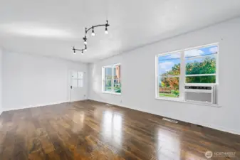 Expansive windows fill the living room with natural light, highlighting the rich wood floors and clean white walls.