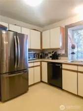 This angle showcases the roomy U-shaped layout, offering generous cabinet storage and ample counter space for cooking and coffee-making alike. The stainless-steel refrigerator and large window add both functionality and natural light, making the kitchen feel bright, practical, and inviting.