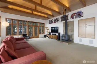 Light-filled living room with soaring ceilings, exposed beams, and a cozy wood stove, designed to capture water views and create a warm, inviting gathering space.