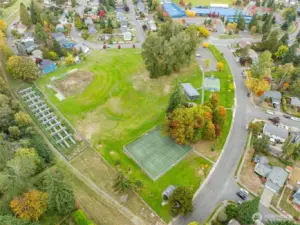 Aerial view of the expansive park shows baseball and tennis courts, a spider web climbing structure, and walking paths throughout. The space is bordered by residential streets and a nearby school.
