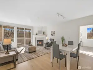 Open living and dining area with gas fireplace and sliding door access to covered deck overlooking wooded greenbelt.
