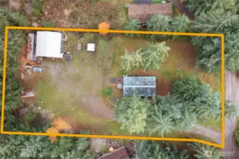 A drone view outlining the full 1.22-acre property, showing the home, the detached shop, and the wide open space between them. The lot is surrounded by tall evergreens for privacy and gives a great sense of the layout, usable areas, and separation between structures. This overhead view highlights how much room there is for parking, projects, and outdoor living.