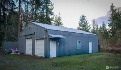 A closer look at the expansive 36x36 shop, featuring two large doors for easy access to vehicles, equipment, or workshop projects. The solid metal construction and wide concrete apron speak to durability and usability. This angle highlights the generous workspace, the storage container alongside the building, and the practical setup that makes this shop a standout feature of the property.
