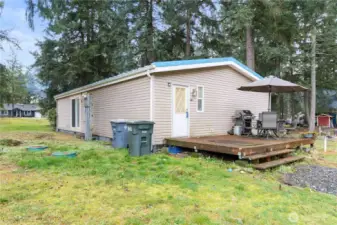 A side view of the home showing the back deck and the open yard surrounding the property. This angle highlights the level outdoor space, the forested backdrop, and the quiet setting. The deck offers plenty of room for outdoor dining and everyday use in a peaceful, natural environment.