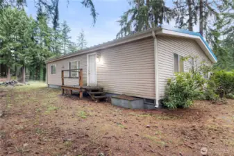 A closer look at the side entry of the home, framed by tall evergreens for added privacy. The natural ground cover and mature landscaping emphasize the wooded setting, while the simple porch provides easy access and a welcoming secondary entrance.