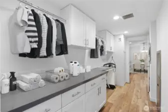 A staged look at the spacious laundry room, featuring abundant counter space, upper cabinets for storage, and a full-length hanging rack. This room is designed for efficiency and organization, with plenty of workspace for folding, sorting, and household tasks.