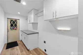 A straightforward view of the laundry room showing the updated cabinetry, long countertop for folding, and built-in hanging racks for easy organization. The space offers practical storage and a functional layout, with direct access to the exterior.