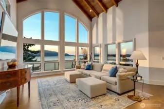 As evening falls, the family room transforms with a warm glow while the windows frame shifting skies and reflections across the water.