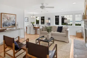 The living room is bright and welcoming, anchored by large windows that fill the space with natural light. A neutral color palette, wood floors, and clean lines create a warm yet modern feel. The layout flows seamlessly into the adjacent dining area, making it ideal for both relaxed living and entertaining.