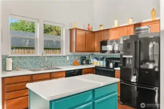 Bright and modern kitchen featuring a spacious island with teal accents, sleek appliances, and a stylish backsplash. Large windows provide plenty of natural light.