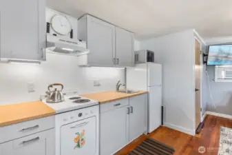 Compact kitchen with white cabinets, laminate butcher block look counters, open shelving, and full-size fridge.