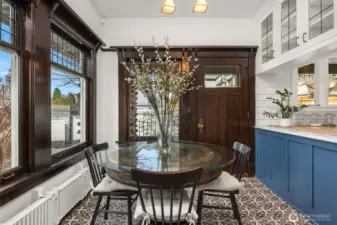A charming breakfast nook blends historic character with modern function. Original wood-trimmed windows, patterned tile floors, and a cozy built-in feel create an inviting space for morning coffee or casual meals, seamlessly connected to the kitchen while overlooking the garden beyond.